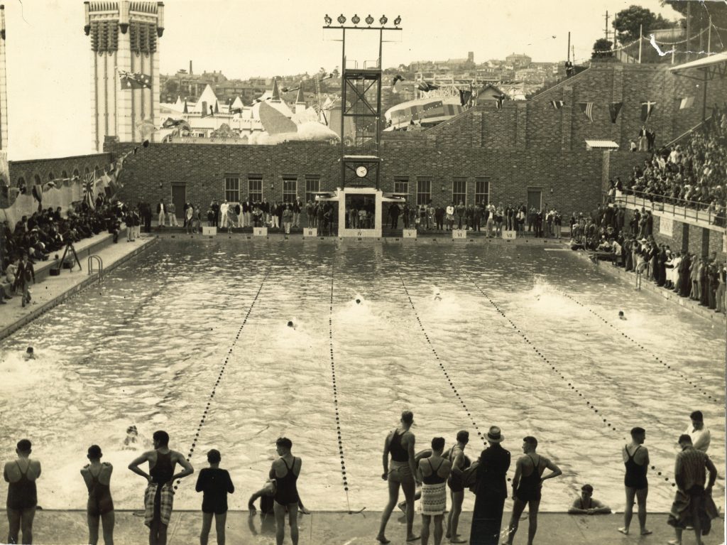 A swimming carnival at North Sydney Olympic Pool in the 1930s, with a diving tower, strings of flags and packed grandstands, and Luna Park on the foreshore behind.