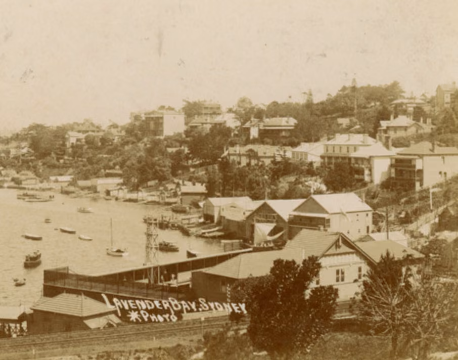 Sepia photograph of Lavender Bay, with the timber Lavender Bay Baths on the shoreline below houses on the wooded hillside and small boats moored offshore.