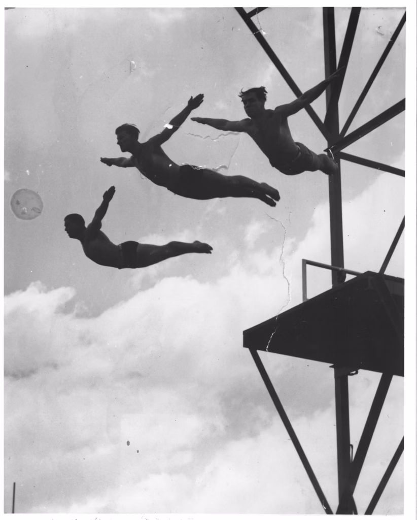 Three divers in mid-flight with arms outstretched after leaping from the high diving platform, photographed from below against a cloudy sky.
