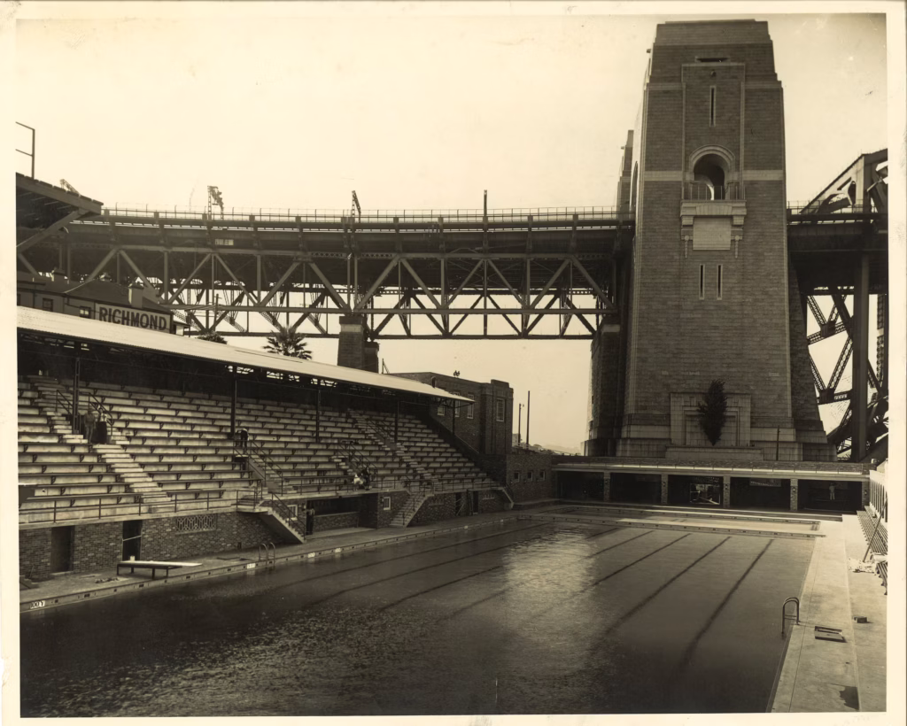 The newly completed North Sydney Olympic Pool, empty of swimmers, with its tiered grandstand along one side and a sandstone pylon of the Sydney Harbour Bridge rising behind it.
