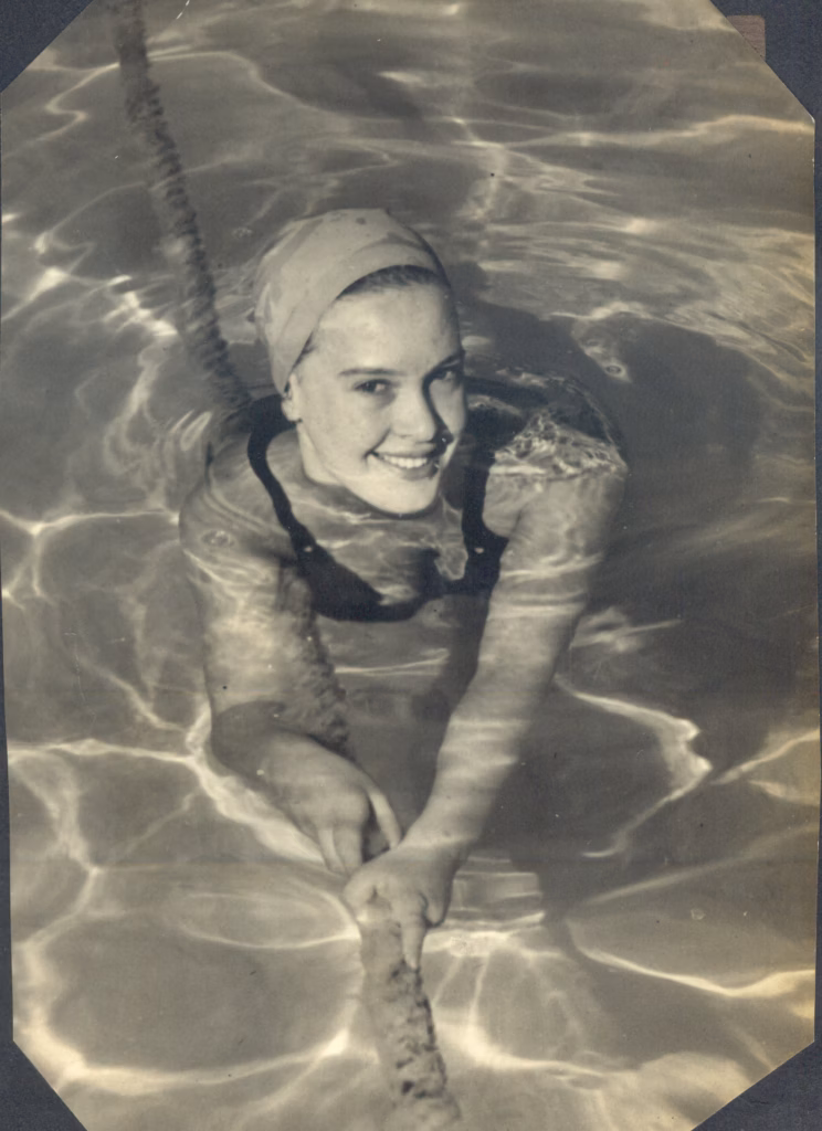 A young woman in a swimming cap smiles up at the camera from the water, holding on to a rope stretched across the pool.