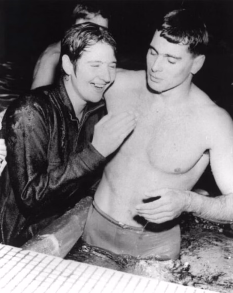 Dawn Fraser, in a wet tracksuit, stands at the pool edge smiling and talking with a swimmer in the water beside her.