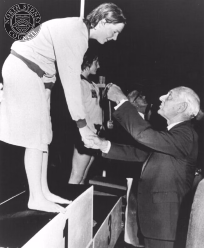Michelle Ford stands on the first-place podium in a tracksuit, smiling and shaking hands as an official reaches up to place a medal around her neck.