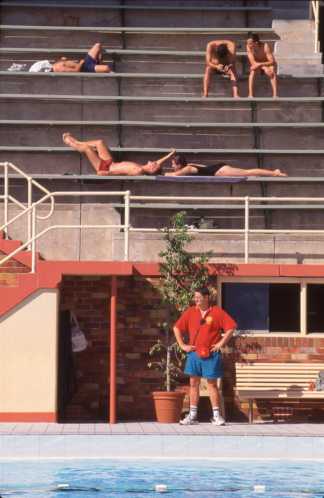 Colour photograph of swimmers sunbathing on the wide concrete grandstand terraces above the pool, with a lifeguard in a red shirt standing on the deck below.