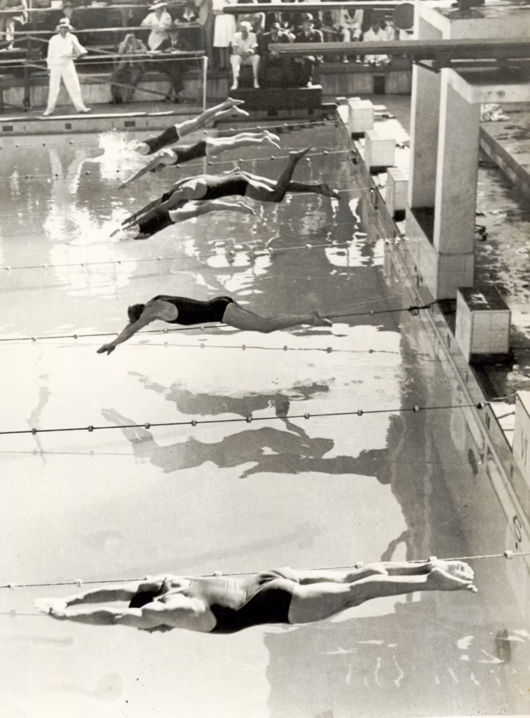 Swimmers pushing off backwards from the wall at the start of a race in the outdoor pool, seen from above with officials watching from the deck.